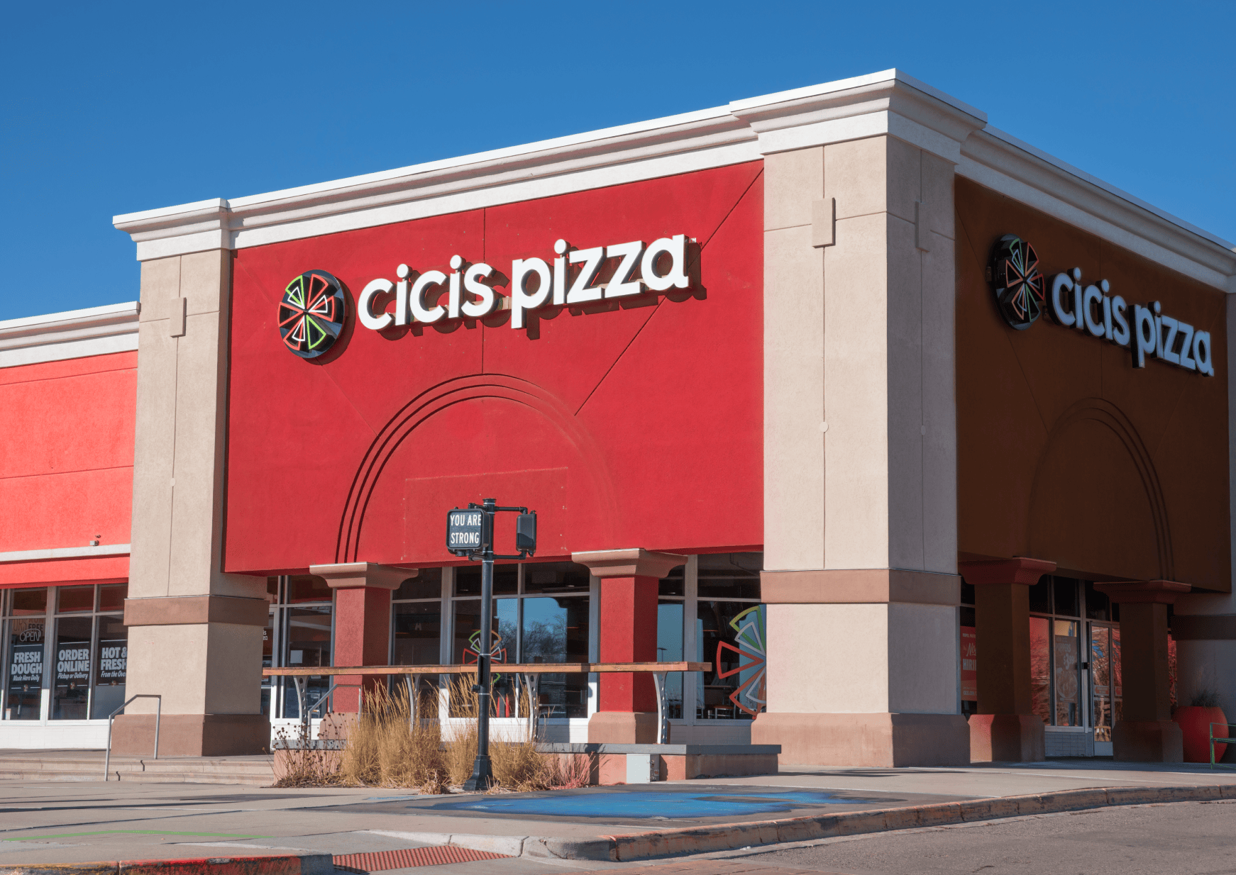 Exterior of a Cicis Pizza restaurant. The square two-story building is bright red with a 'Cicis Pizza' sign and pie spark logo over the two adjacent entrances.
